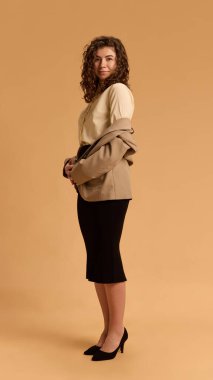 Kendine güven. Formalite giymiş genç ve çekici bir kadının bej renkli stüdyo arkaplanına zarif bir şekilde poz verdiği uzun bir fotoğraf. İnsani duygular, güzellik, iş, moda ve tarz kavramı.