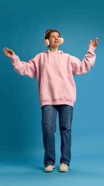 Pembe kapüşonlu neşeli genç bir kadın, şarkı söyleyip dans ediyor, kulaklıkla mavi stüdyo arka planında müzik dinlerken ellerini kaldırıyor. İnsani duygular, güzellik, moda ve tarz kavramı.