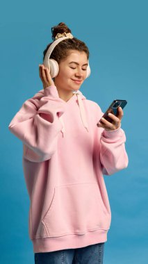 Gündelik giyinmiş genç, güzel bir kadın, telefondaki şarkı listesini seçiyor kulaklıkla mavi stüdyo arka planında dinlemek için. İnsani duygular, güzellik, moda ve tarz kavramı.