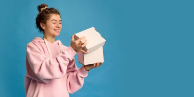 Gündelik giyinmiş genç güzel bir kadın mavi stüdyo arka planına karşı tuhaf ve şaşırmış bir ifadeyle kutuyu açıyor. Uzayı kopyala İnsani duygular, güzellik, moda ve tarz kavramı.