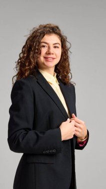 Kıvırcık, resmi giyinmiş genç bir kadının dikey portresi. Minimalist stüdyo arka planına gülümseyen bir şekilde kameraya bakıyor. İnsani duygular, güzellik, iş, moda ve tarz kavramı.