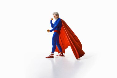 Kır saçlı, süper kahraman kostümlü, kırmızı pelerinli, dambılı beyaz stüdyo arka planında. Sağlıklı ve sportif yaşam tarzının tanıtımı. Spor anlayışı, egzersiz, vücut bakımı