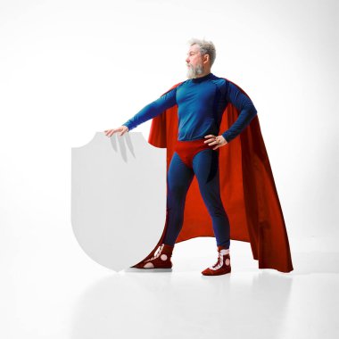 Beyaz stüdyo arka planında beyaz kalkanla duran kırmızı pelerinli süper kahraman kostümlü özgüvenli bir adamın tam boy portresi. Sigorta, koruma, güvenlik, koruma kavramı