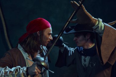 İki odaklanmış genç adam, klasik korsan kostümleri giymiş kılıçlarla yoğun bir düelloya tutuştular, yakın durdular ve koyu dokulu arka planda birbirlerine baktılar. Tarih kavramı