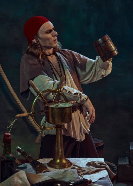 Genç bir adam, kırmızı bandanalı bir korsan, gemi tekerine yaslanmış, kara arka planda deniz aletleriyle içki içerek ahşap bardağa bakıyor. Tarih kavramı, macera, keşif, yaşam tarzı