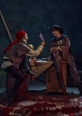 İki adam, klasik kostümlü korsanlar, kart oyunu oynuyorlar, etrafı karanlık arka plana karşı dünya, teleskop ve klasik harita gibi aksesuarlarla çevrili. Kumar, macera, oyun kavramı