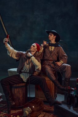 İki adam, bandana ve şapka takmış korsanlar, oturuyorlar ve arka planda akıllı telefonla selfie çekiyorlar. Cesaret. Tarih kavramı, çağların karşılaştırması, eğlence.