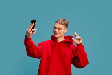 Genç bir adamın portresi, normal kırmızı kapüşonlu, akıllı telefonuyla poz veren, mavi stüdyo arka planına karşı selfie çeken. İnsan duyguları kavramı, güzellik, kendini ifade etme, sosyal medya.