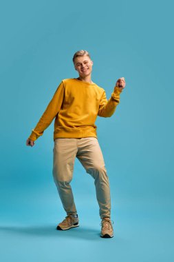 Dinamik, genç bir adamın tam vücut fotoğrafı, şık kıyafetler giymiş, dans ediyor, mavi stüdyo arka planına karşı enerjik bir şekilde el sallıyor. İnsan duyguları, güzellik ve moda kavramı, kendini ifade etme..