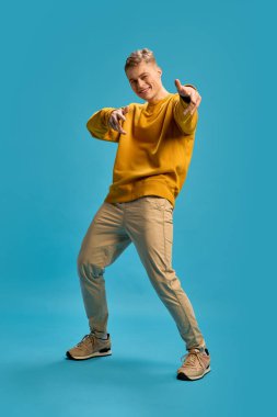 Adam, neşeli bir gülümsemeyle, hafifçe eğilip, mavi stüdyo arka planına karşı enerjik jestler yaparak ileri işaret ediyor. İnsan duyguları kavramı, güzellik, kendini ifade etme.