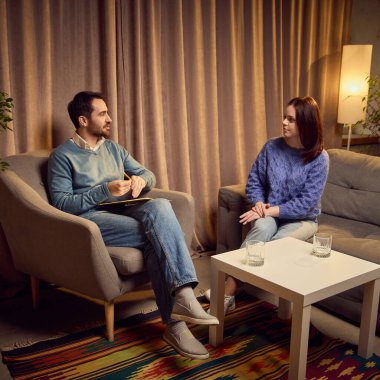 Mavi kazaklı bir erkek psikolog, elinde not defteri, rahat kıyafetler giyen genç bir kadınla terapi seansında, rahat bir odada sandalyelerde oturuyor. Psikoloji kavramı, yardım, terapi