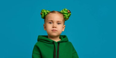 Güzel bir çocuğun portresi, yeşil kapüşonlu bir kız ve örülmüş çörekler, sakin bir yüzle poz veriyor, mavi stüdyonun arka planına bakıyor. Çocukluk kavramı, duygular, yaşam tarzı