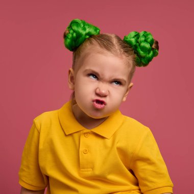 Çocuk, sinirli suratlı, yeşil örülmüş çörekler saç stili, sarı gömlekli, pembe stüdyo geçmişli bir kız. Komik surat. Çocukluk kavramı, duygular, yaşam tarzı