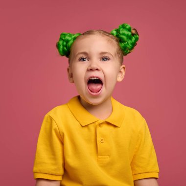 Çocuk, kız heyecanını açık ağızla ifade ediyor, sarı gömlek giyiyor, yeşil örülmüş saç stili pembe stüdyo arka planına karşı. Neşe. Çocukluk kavramı, duygular, yaşam tarzı