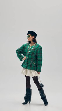 Ekose yeşil ceket, inci kolye, beyaz etek, topuklu çizmeler, minimalist stüdyo geçmişine karşı bere ve güneş gözlüğü takan zarif genç bir kadın. İş anlayışı, moda eğilimleri, güzellik, tarz