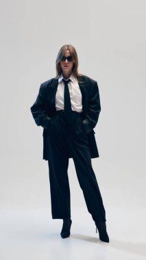 Güneş gözlüklü genç bir kadın siyah ceket ve pantolon, topuklu botlar, beyaz gömlek ve kravat giyer, stüdyo arka planında şık ama klasik bir kıyafet giyer. İş anlayışı, moda eğilimleri