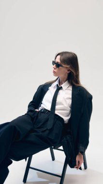 Güneş gözlüklü, rahat bir pozisyonda oturan, siyah kostümlü, beyaz gömlekli ve kravatlı şık bir şekilde poz veren kendine güvenen bir iş kadını. İş, moda konsepti