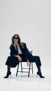 Güneş gözlüklü bir iş kadını, kendine güvenen bir pozisyonda oturuyor, siyah kostümlü, beyaz gömlekli ve stüdyo arka planında kravat takıyor. İş anlayışı, moda eğilimleri