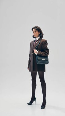 Siyah taytlı, kendine güvenen, şık bir genç kadın, kahverengi büyük beden ceket, kravat ve stüdyo arka planına karşı zarif poz veren küçük bir çanta. İş anlayışı, moda eğilimleri, güzellik, tarz