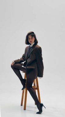 Siyah taytlı modaya uygun bir kadın ve kravatlı kahverengi bir spor ceket, kendine güvenen bir pozda oturuyor ve gri stüdyo arka planında poz veriyor. İş anlayışı, moda eğilimleri, tarz