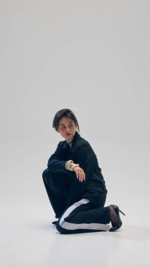 Siyah kostümlü, spor pantolonlu, spor ceketli, inci bilezik ve topuklu ayakkabılı stüdyo geçmişine karşı poz veren zarif bir genç kadın. İş anlayışı, moda eğilimleri, güzellik, tarz