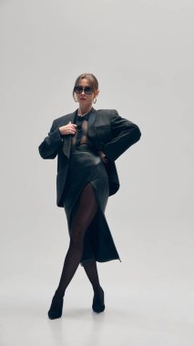 Siyahlar içinde kendine güvenen bir kadın, büyük beden ceket, bluz ve deri etek, gri stüdyo arka planına karşı elinde çantayla güvenle poz veriyor. İş anlayışı, moda eğilimleri, güzellik, tarz