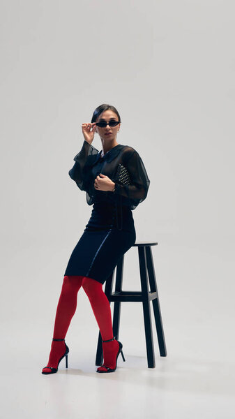 Graceful young woman in black blouse, skirt, red tights, heels and sunglasses posing on chair against grey studio background. Concept of business, fashion trends, beauty, style