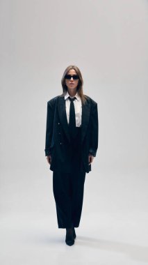 Siyah kostümlü, büyük beden ceket, pantolon, beyaz gömlek ve kravatlı, güneş gözlükleriyle stüdyo arka planında yürüyen kendinden emin genç bir iş kadını. İş anlayışı, moda eğilimleri