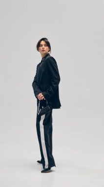 Siyah takım elbiseli, ceketli ve küçük çantalı güzel bir genç kadının stüdyo arka planında poz verirken tam boy portresi. İş anlayışı, moda eğilimleri, güzellik, tarz