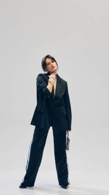 Siyah takım elbiseli güzel bir genç kadının tam boy portresi, pantolon, ceket, küçük çanta ve gri stüdyo arka planına poz veren inci kolye. İş anlayışı, moda eğilimleri, güzellik, tarz