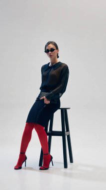 Siyah bluz, etek, kırmızı tayt, topuklu ayakkabı ve güneş gözlüğü giyen zarif bir kadın gri stüdyo arka planına karşı koltukta güven içinde poz veriyor. İş anlayışı, moda eğilimleri, güzellik, tarz