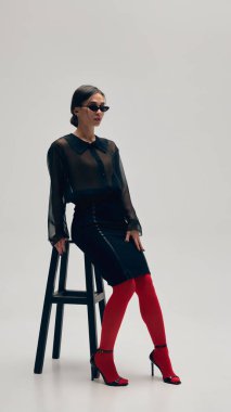 Siyah bluz, etek, kırmızı tayt, topuklu ayakkabı ve güneş gözlüğü giymiş zarif genç bir kadın gri stüdyo arka planına karşı sandalyede poz veriyor. İş anlayışı, moda eğilimleri, güzellik, tarz