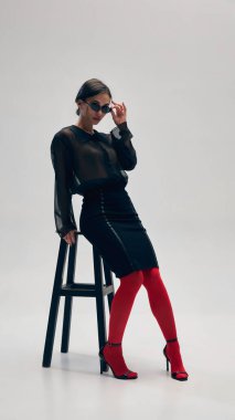 Siyah bluz, etek, kırmızı tayt, topuklu ayakkabı ve güneş gözlüğü giyen zarif bir kadın gri stüdyo arka planına karşı koltukta güven içinde poz veriyor. İş anlayışı, moda eğilimleri, güzellik, tarz