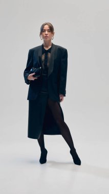 Tamamen siyah kıyafetli güzel bir iş kadını, büyük beden ceket, bluz ve etek, gri stüdyo arka planına karşı elinde çantayla güvenle ayakta duruyor. İş stili kavramı, moda eğilimleri