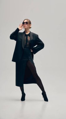 Siyahlar içinde kendine güvenen bir kadın, büyük beden ceket, bluz ve deri etek, gri stüdyo arka planına karşı elinde çantayla güvenle poz veriyor. İş anlayışı, moda eğilimleri, güzellik, tarz