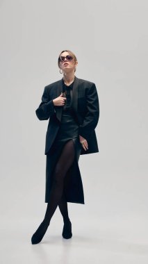 Güneş gözlüklü, kendine güvenen zarif bir kadın, büyük beden ceket, bluz ve deri etek, açık gri stüdyo arka planında poz veriyor. İş anlayışı, moda eğilimleri, güzellik, tarz