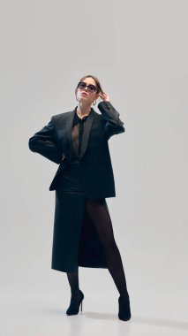 Tamamen siyah kıyafetli iş kadını portresi, büyük beden ceket, bluz, deri etek ve güneş gözlüğü açık gri stüdyo arka planına karşı güvenle poz veriyor. İş anlayışı, moda eğilimleri