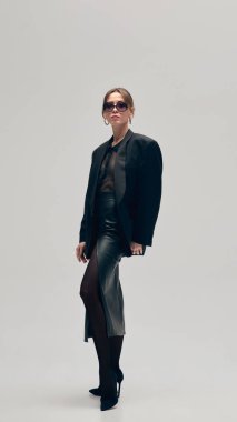 Tamamen siyah kıyafetli bir portre iş kadını, büyük beden ceket, bluz, deri etek ve güneş gözlüğü açık gri stüdyo arka planına karşı güvenle poz veriyor. İş anlayışı, moda eğilimleri, tarz