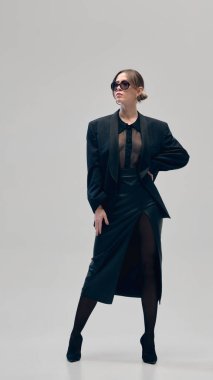 Tamamen siyah kıyafetli bir portre iş kadını, büyük beden ceket, bluz, deri etek ve güneş gözlüğü açık gri stüdyo arka planına karşı güvenle poz veriyor. İş anlayışı, moda eğilimleri