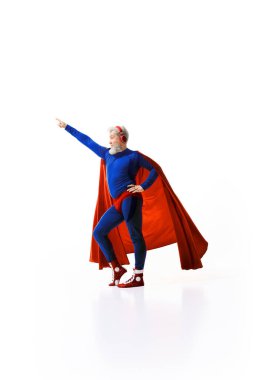 Gri saçlı süper kahraman kostümlü, kırmızı pelerinli, kulaklıkla müzik dinleyen ve beyaz stüdyo arka planına yumruk kaldıran bir adamın tam boy görüntüsü. Kahramanlık kavramı, fantezi, yaşam tarzı