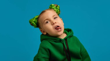 Anaokulu çocuğunun portresi, yeşil kapüşonlu duygusal kız ve güzel popolu saç stili, surat asma, mavi stüdyo arka planına karşı suratını buruşturma. Can sıkıntısı. Çocukluk kavramı, duygular, yaşam tarzı