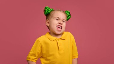 Duygusal anaokulu kızı portresi, sarı tişörtlü çocuk ve örülmüş çörekler, komik suratlar yapmak, suratını buruşturmak, pembe stüdyo geçmişine karşı oynamak. Çocukluk kavramı, duygular, yaşam tarzı