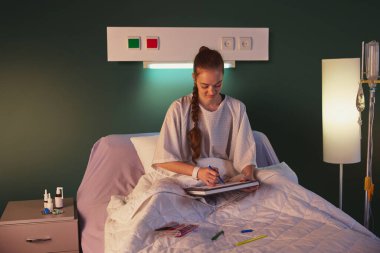 20 'li yaşlarda genç bir kız hastane önlüğüyle yatakta oturuyor, seruma benzer tıbbi ekipmanlarla, ve akşamları çizim yapıyor. Olumlu iyileşme, modern klinik. Sağlık, ilaç, tedavi kavramı