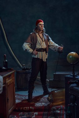 Ortaçağ korsanı gibi giyinmiş kırmızı bandanalı genç bir adamın portresi koyu renkli arka planda Rapier ile duruyor. Cesaret ve isyan kavramı, tarih, keşif.