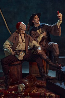 İki adam, bandana ve şapka takmış ortaçağ korsanları, karanlık arka planda oturup akıllı telefonlu selfie çekiyorlar. Cesaret. Sosyal ortam kavramı, dijital ağ kurma, dönemlerin karşılaştırması