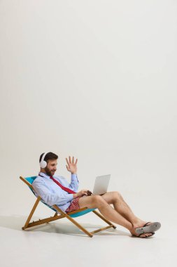 Resmi gömlekli, kravatlı, şortlu, kulaklıklı plaj sandalyesinde oturan iş görüşmeleri sırasında dizüstü bilgisayara el sallayan bir iş adamı. Uzaktan çalışma, tatil, iş-yaşam dengesi kavramı