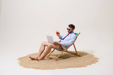 Plaj sandalyesinde oturan, dizüstü bilgisayarda çalışan, kokteyl tutan, güneş gözlüğü takan, resmi gömlek giyen, kravat takan, mayo giyen ve kumda çıplak ayaklı bir adam. Uzaktan çalışma, tatil, seyahat, iş hayatı dengesi kavramı