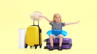 Heyecanlı çocuk, bavulun üzerinde oturan kız, kolları tamamen açık, şişirilebilir yüzme yüzüğü takıyor, sarı stüdyo arka planında tatile gitmeyi hayal ediyor. Çocukluk kavramı, tatil, seyahat, turizm