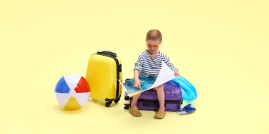 Çizgili gömlekli ve turuncu güneş gözlüklü bir çocuk sarı stüdyo arka planına bakarak haritaya bakıyor. Yaz deniz gezisi. Çocukluk kavramı, tatil, seyahat, turizm