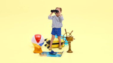 Dürbünle bakan bir çocuk. Gemi tekerleği, şişirilebilir halka da dahil olmak üzere seyahat aksesuarlarıyla çevrili. Sarı stüdyo arka planında basılı harita. Çocukluk, tatil, seyahat, turizm